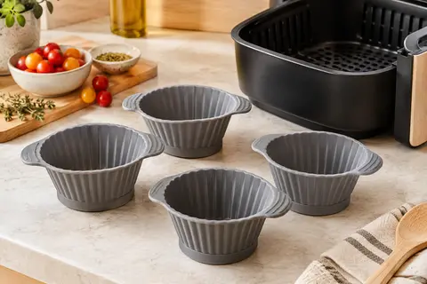 Quatre moules à muffins gris en silicone avec poignées, posés près d’un panier de friteuse à air.
