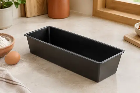 Moule à cake noir en acier revêtu posé sur un plan de travail clair avec quelques ingrédients de pâtisserie.